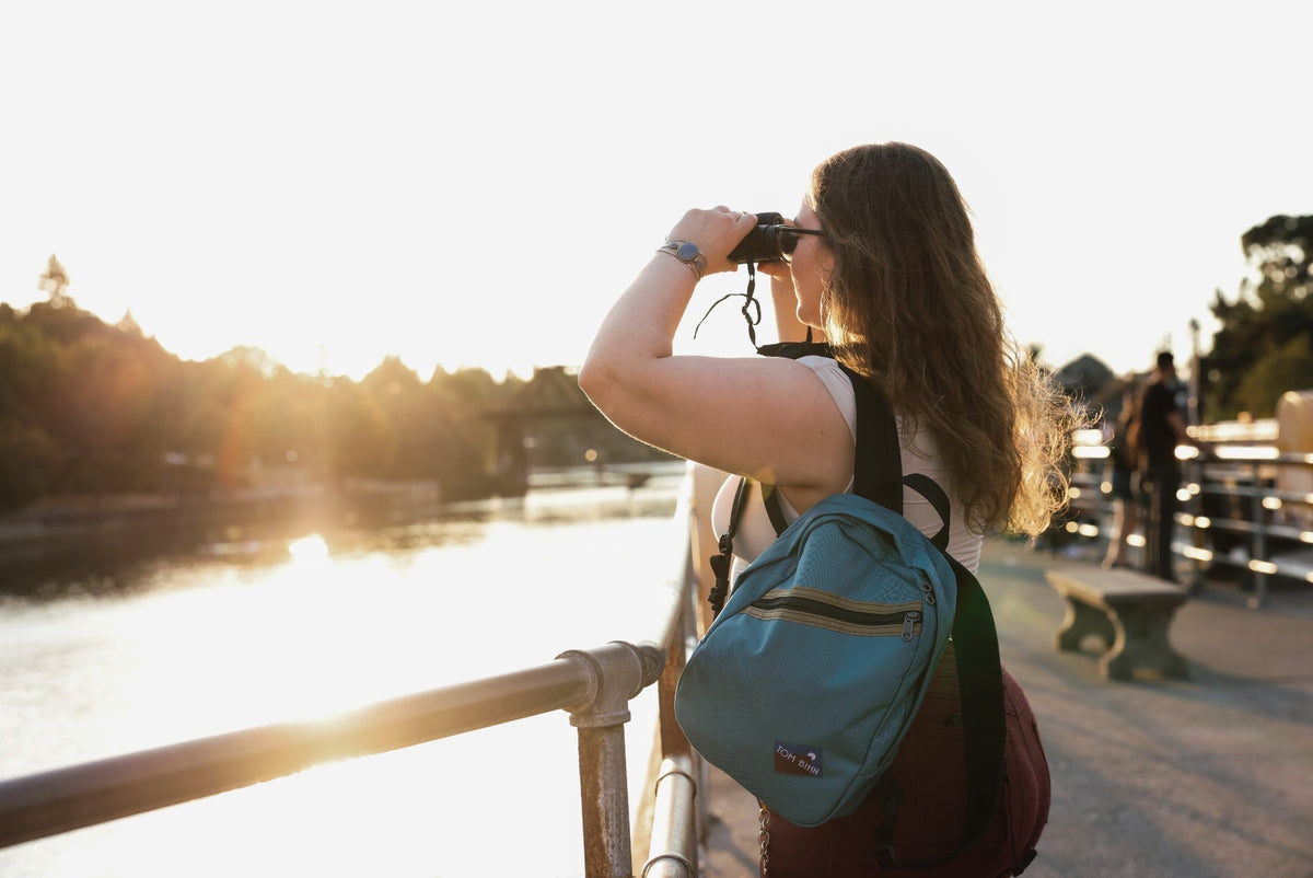 Person wearing a Zeitgeist on their shoulder, using binoculars looking out over water.