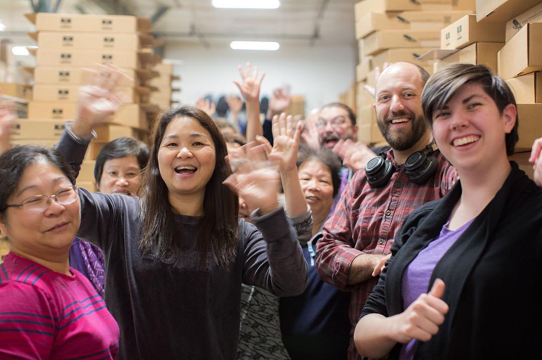 Sau Ling, Zhong, Mike, and Kat. Everyone's waving and smiling and there's huge stacks of shipping boxes behind them.