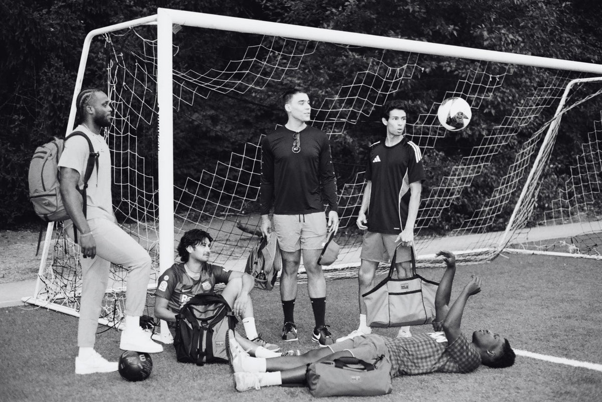 Black and white image of five people standing near a soccer goal holding TOM BIHN bags. From left: Synik 30, Brain Bag, Old School Carry All, Yeoman Duffel, and Monster Truck.