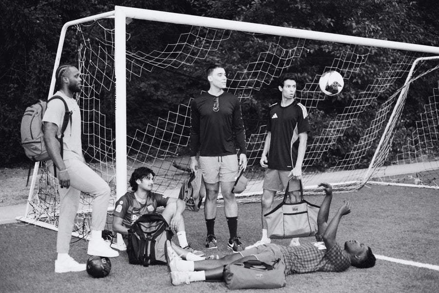 Black and white image of five people standing near a soccer goal holding TOM BIHN bags. From left: Synik 30, Brain Bag, Old School Carry All, Yeoman Duffel, and Monster Truck.