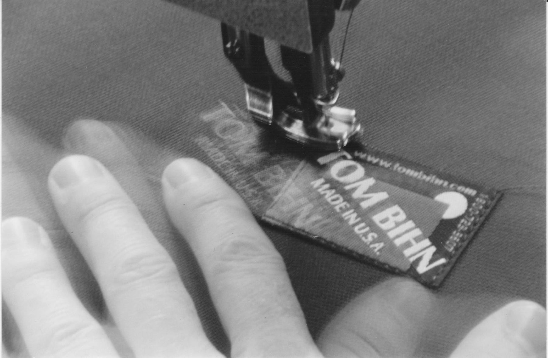 The photo shows Tom's hands blurred as he sews a TOM BIHN label onto a bag.