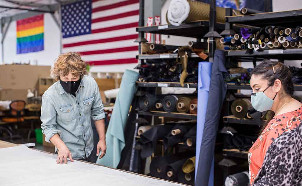 Nik and Irma at the Seattle factory discussing a marker. There's a rainbow flag and an American flag in the background.