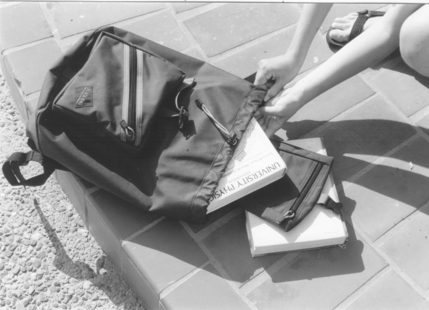 A basic book bag on the ground: a person is pulling books out of the main compartment.