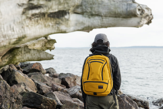 A person wearing backpack standing on rocks by a body of water.