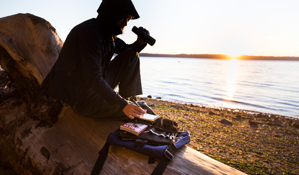 visible lifestyle^^Person sitting on a log at the beach, holding a pair of binoculars and taking notes. The Large Cafe Bag with its Freudian Slip sits next to them.