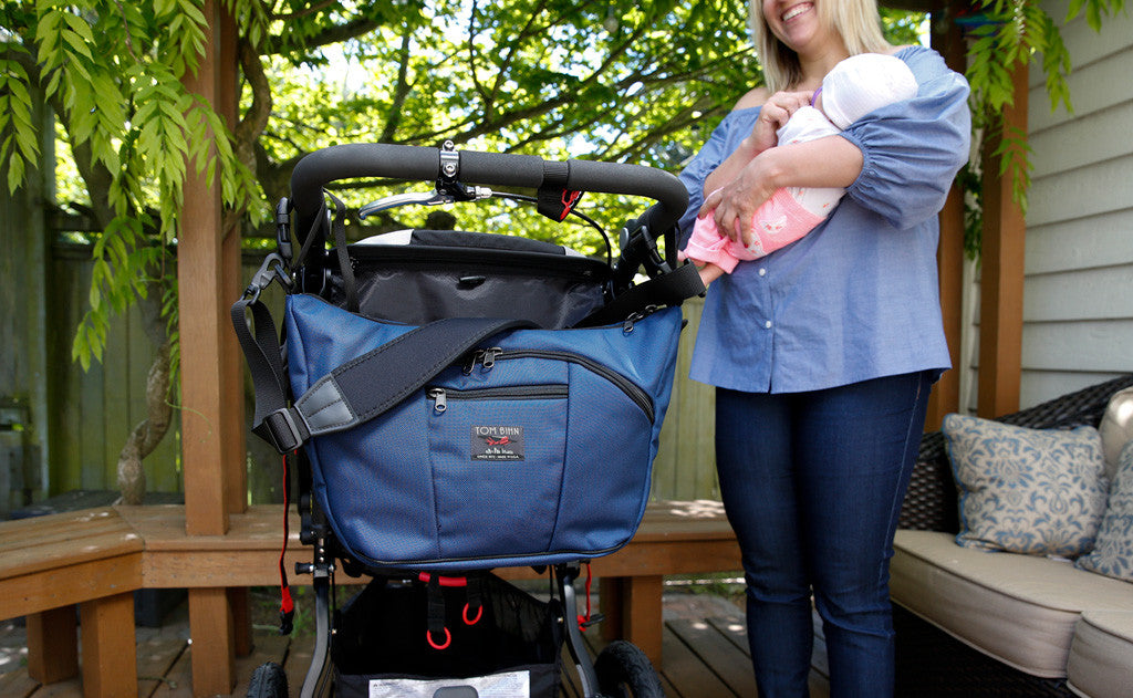 lifestyle visible^^A person holding a baby beside a Deep Blue (dark blue) 525 Ballistic Parental Unit connected to the push handle of a stroller using the stroller/wheelchair adapter straps.