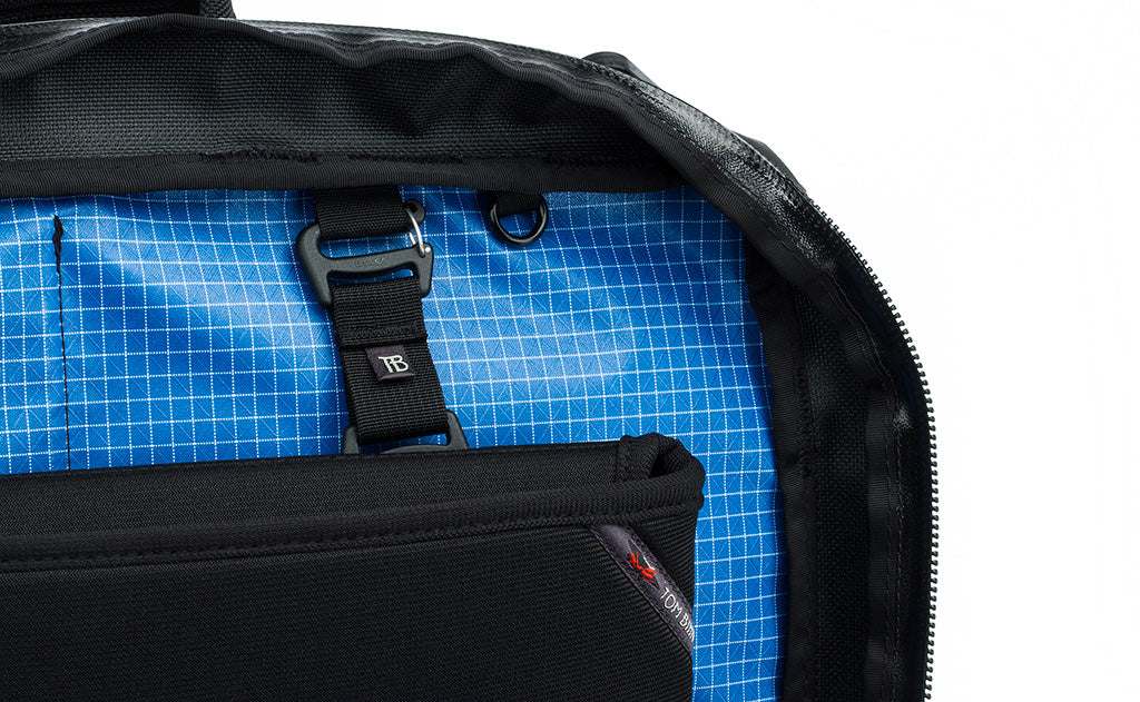 A close shot of the rear main pocket of a Black 525 Ballistic exterior Western Flyer opened to reveal an Island (blue) 200 Halcyon interior with a Cache connected to the nylon loops sewn into the interior seam and a nearby o-ring.
