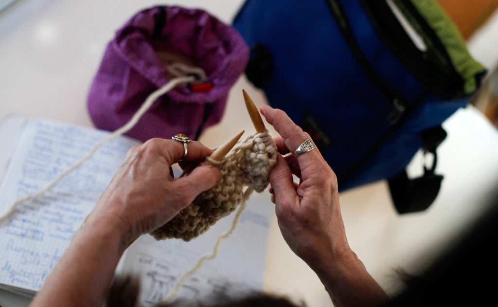 ^^A person knitting with her Maker's Bag on the table next to their Yarn Stuff Sack and notes.