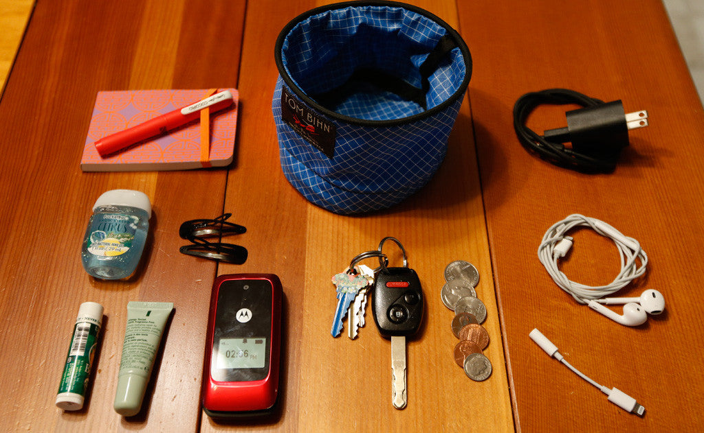 Small Travel Tray surrounded by various accoutrements, a charge cable, earbuds, a dongle, a bit of change, a pair of keys, a notepad and pen, lotion, chapstick, hair-clips and a flip phone