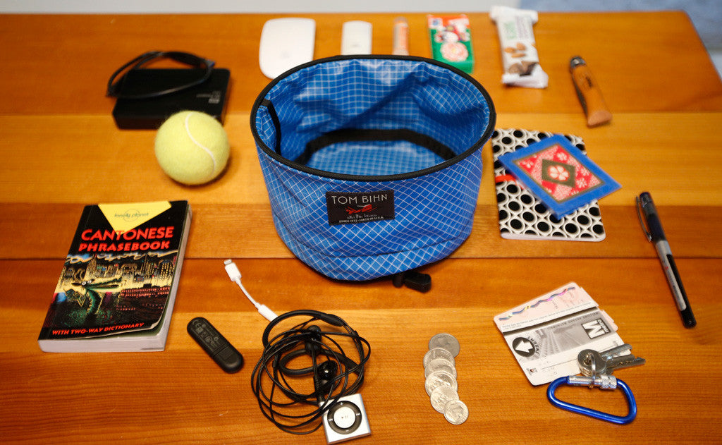 ^A Large Travel Tray surrounded by various accoutrements: a Cantonese Phrasebook, a USB keydrive, a charging battery, a dongle, a pair of earbuds and MP3 player, a bit of change, a carabiner and key, a Metrocard, a pen, a notepad, a tube of lotion, some snacks, and a tennis ball.
