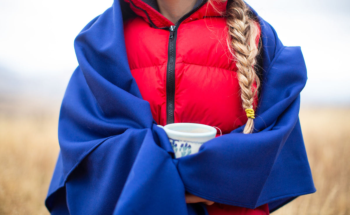 lifestyle visible^^A person wearing a bright red down vest, standing outdoors on an overcast winter day; their hair is in a long braid and they are holding a mug of hot tea. The Shepherd's Wool in Neela Blue is wrapped around their shoulders and hands for warmth.
