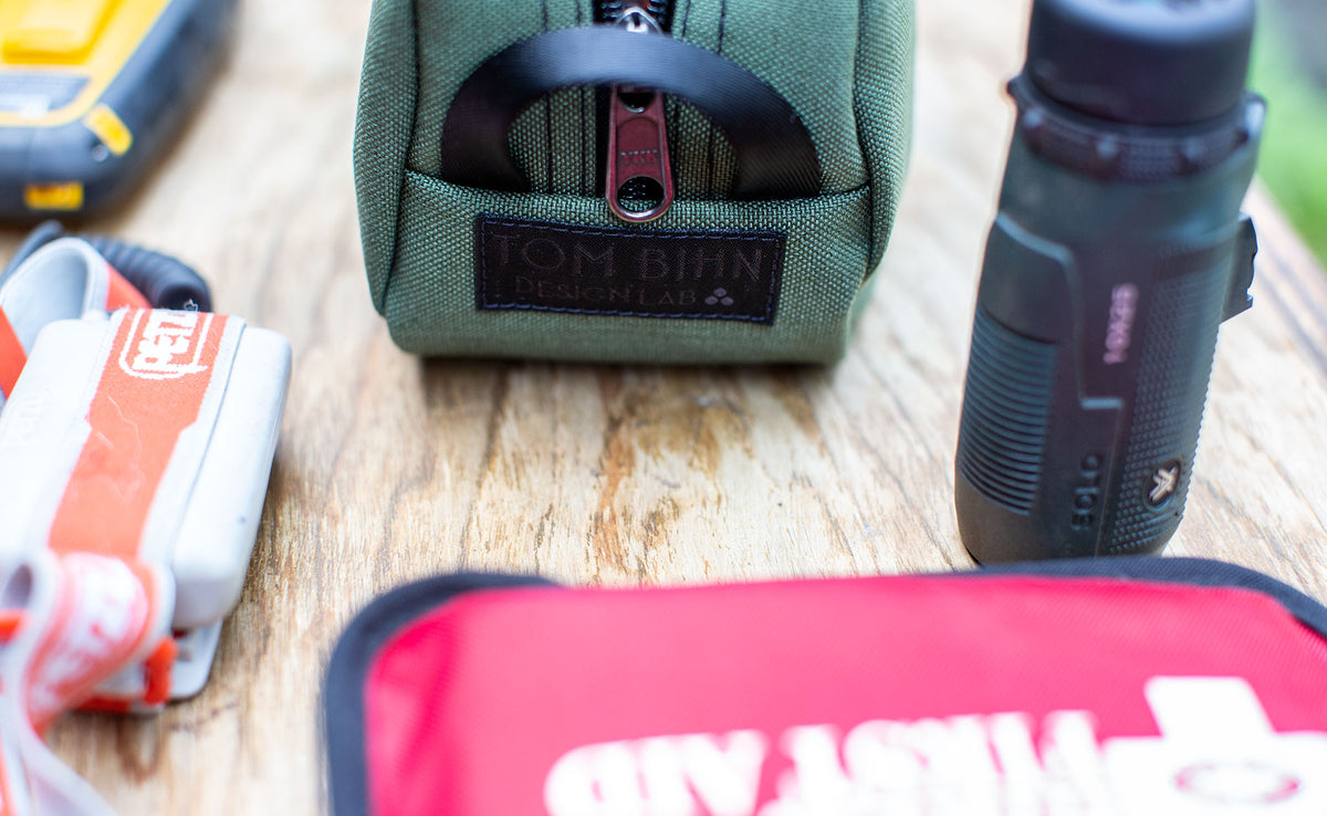 detail visible^^ A Boreal Grab Bag showing the TOM BIHN Design Lab label on a picnic table with a headlamp, First Aid Kit, and flashlight. 