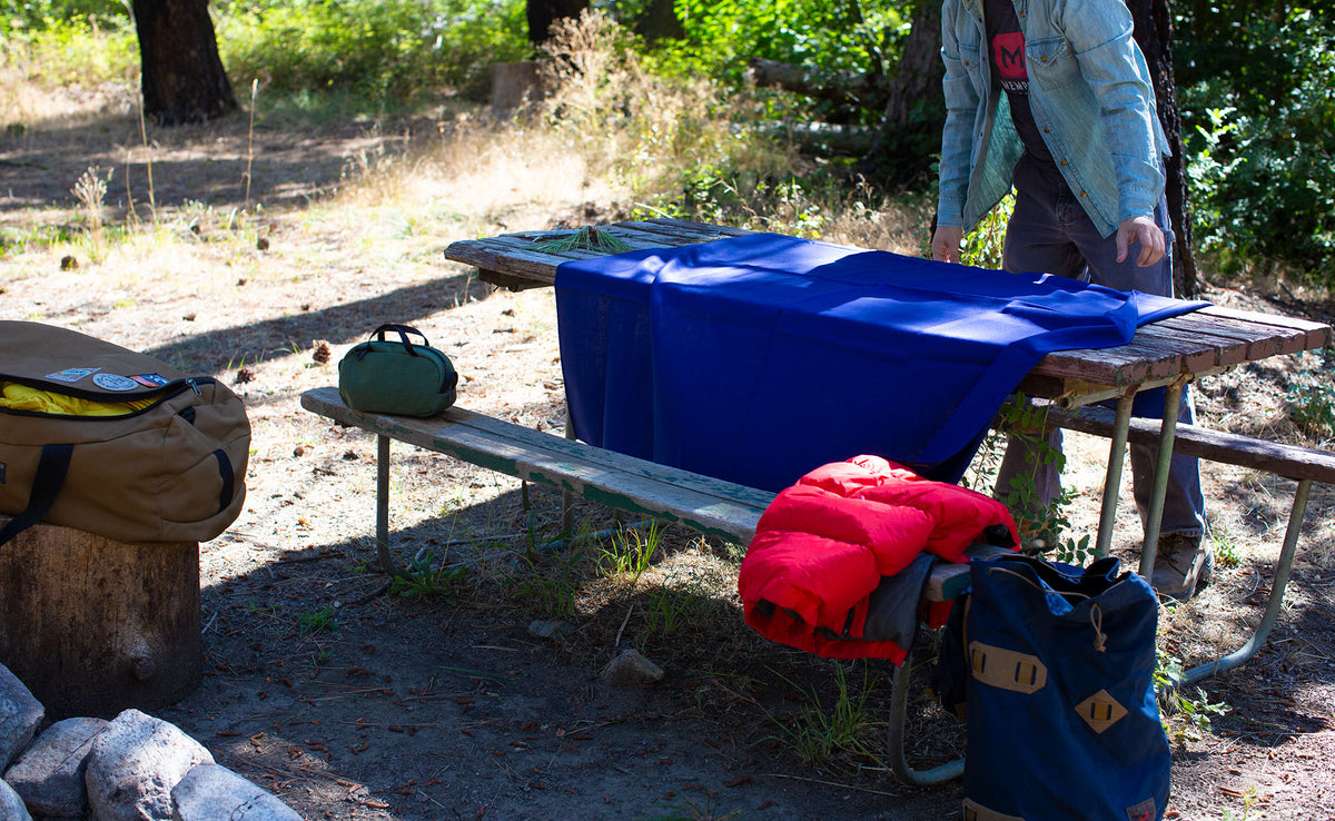 lifestyle visible^^ A Large Shepherd's Wool Utility Cloth in Neela Blue spread out onto a picnic table at a campsite.