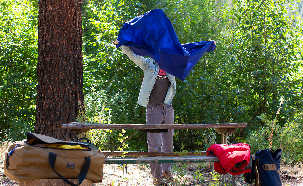 lifestyle visible^^ A Large Shepherd's Wool Utility Cloth in Neela Blue being shaken into the air to spread as a table cloth on a picnic table at a campsite. 