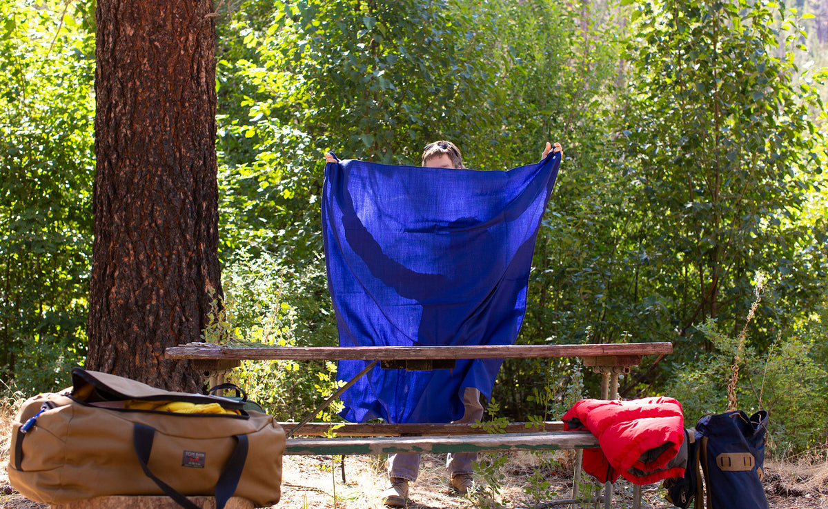lifestyle visible^^ A Large Shepherd's Wool Utility Cloth in Neela Blue being shaken into the air to spread as a table cloth on a picnic table at a campsite.