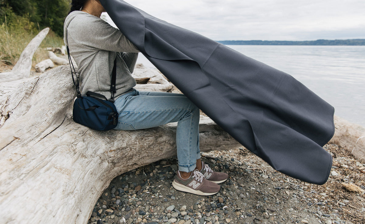 lifestyle visible^^ A person sitting on a log at the beach getting ready to lay a Large Shepherd's Wool Utility Cloth in Matter Grey on their lap. 