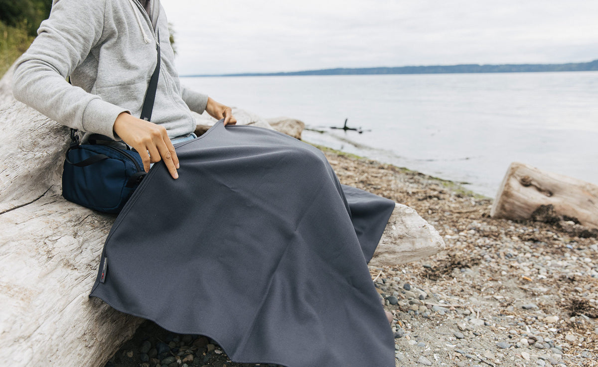 lifestyle visible^^ A person sitting on a log at the beach with a Large Shepherd's Wool Utility Cloth in Matter Grey on their lap.