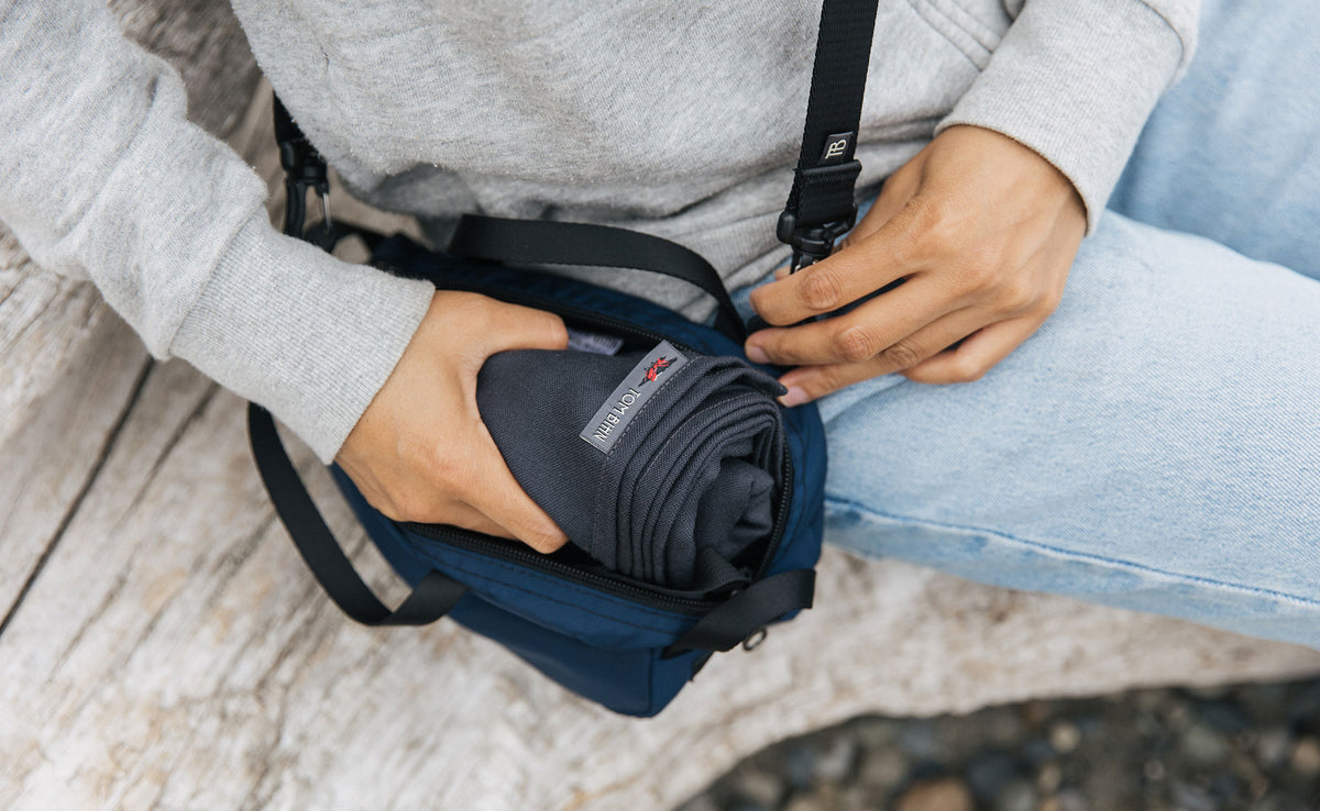 lifestyle visible^^ A person sitting on a log  with a Large Shepherd's Wool Utility Cloth in Matter Grey rolled and being stuffed into a Navy Parapack Grab Bag.