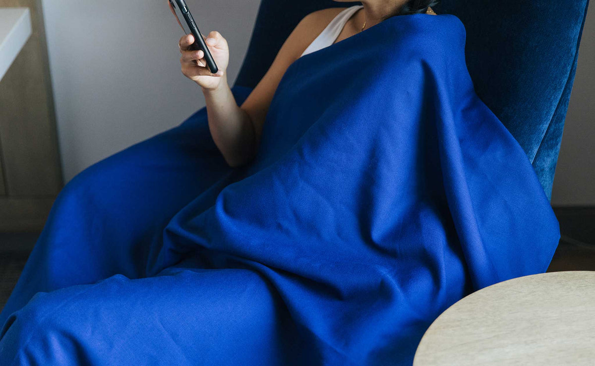 detail visible^^ A Large Shepherd's Wool Utility Cloth in Neela Blue being worn as a blanket while a person rests in a blue velvet chair reading something on their phone.