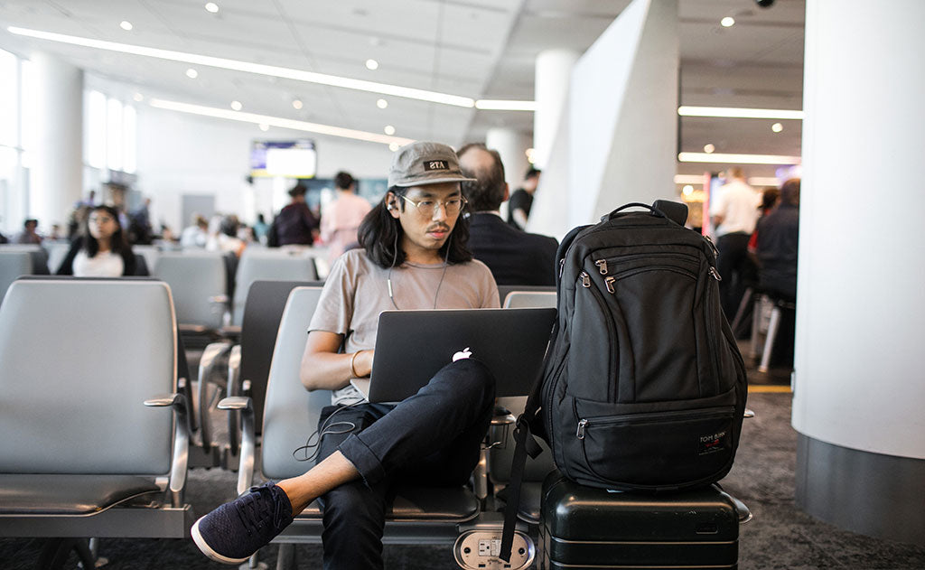 visible lifestyle^^A person working on their laptop on an airport while their Synik 30 sits on their roll-aboard luggage.